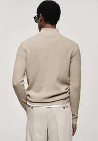 Beżowy sweter o prążkowanej strukturze, z długimi rękawami i wysokim kołnierzem, zestawiony z jasnymi spodniami z guzikami po bokach w pasie.