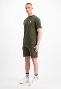 Olijfgroene t-shirt met een logo op de borst, gecombineerd met bijpassende shorts. Het model draagt witte sneakers en gestreepte zwarte sokken.