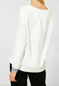 Camisa de manga larga blanca con textura acanalada. Presenta un escote redondo, aberturas laterales y puños en azul claro con texto impreso.