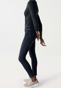 Schmale dunkelblaue Jeans aus Denim mit hoher Taille, ausgestattet mit Gesäßtaschen und dezenten Stickdetails, kombiniert mit einem strukturierten dunklen Oberteil und hellen Sneakern.