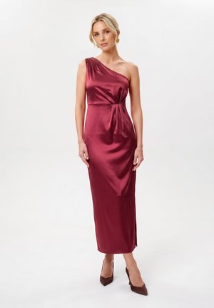 Femme aux cheveux blonds portant une robe en satin bordeaux à une épaule, debout sur un fond blanc, avec des talons foncés et des boucles d'oreilles dorées.