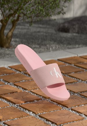 Roze Calvin Klein schuifsandalen met een getextureerde band en een wit logo, rustend op een geordende houten en grindvloer buiten, vlakbij een boom.