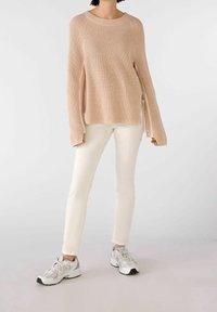 Vaaleanpunainen neulepaita, jossa on pyöreä kaula-aukko ja sivuhalkiot, yhdistettynä valkoisiin slim-fit-farkkuihin ja harmaisiin lenkkareihin, joissa on mustia yksityiskohtia.