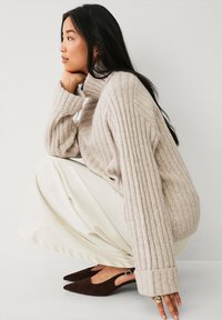 Beige gerippter Rollkragenpullover mit langen Ärmeln, kombiniert mit cremefarbenen weiter geschnittenen Hosen und dunkelbraunen Slingback-Schuhen.