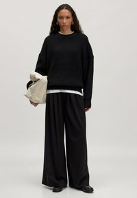 Zwarte oversized gebreide trui met verlaagde schouders, gecombineerd met losse zwarte broek en een crème-kleurige tas. Gladde textuur en relaxte pasvorm.