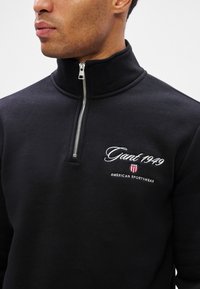Muž v čiernom pulóvri s krátkym zipsom, na ktorom je vyšívaný nápis "Gant 1949" a logo American Sportswear na ľavej hrudi.