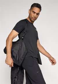 Muž v černém tričku a kalhotách, držící černou bundu přes rameno, dívající se dolů na jednobarevném světle pozadí.