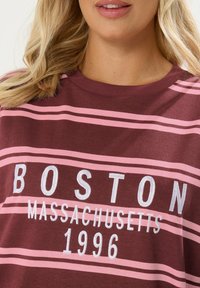 Blondynka w bordo i różowej koszulce w paski z białym napisem "Boston Massachusetts 1996."