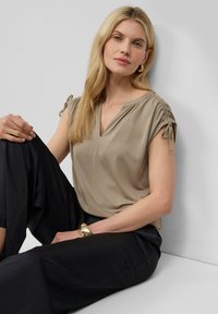 Haut beige à manches courtes avec un col en V, détails froncés aux épaules et texture douce, associé à un pantalon noir ample. Accent d'un bracelet en or.