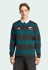 Chemise à manches longues de style rugby avec des rayures horizontales vert foncé et noires, col blanc, logo brodé et accent à trois rayures sur la manche.