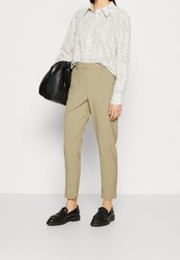 Witte blouse met zwart gespikkeld patroon, hoog-waisted beige broek en zwarte loafers, aangevuld met een grote zwarte handtas.