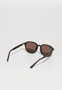 Lunettes de soleil teintées marron avec des verres ronds, monture en plastique marron foncé, accents en métal sur les charnières et logo de la marque gravé sur le côté.