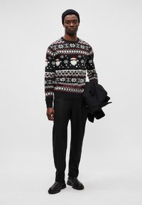 Sweater de lã preta com padrões festivos, incluindo rostos de Pai Natal e flocos de neve, combinado com calças pretas e um casaco na mão.