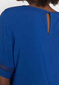 Top blu con maniche corte, dettaglio in pizzo, scollatura rotonda e chiusura con piccolo bottone sul retro con design a occhiello.