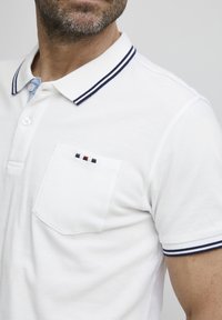 Hvit polo skjorte med blå underkrage, marineblå stripete krage, og en brystlomme med tre små fargede striper (rød, marineblå, hvit).