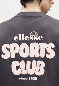Άτομο που φορούσε σκούρο φούτερ με μεγάλο λευκό κείμενο "ellesse SPORTS CLUB since 1959" στην πλάτη, το κεφάλι γυρισμένο ελαφρώς προς τα πλάγια.