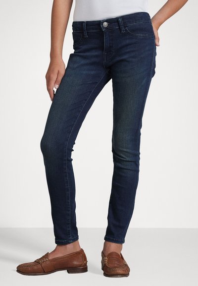 Vaqueros ajustados de color azul oscuro con una textura de denim suave, diseño de cinco bolsillos y cierre de botón plateado en la cintura. Llevados con mocasines marrones.