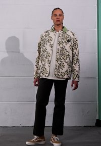 Floral-print jacket in cream with green collar, layered over a white shirt. Paired with dark pants and brown-and-white sneakers.