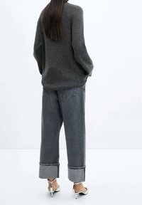 Persoon met lang donker haar, gekleed in een donkergrijze trui, wijd omgeslagen grijze spijkerbroek en witte hoge hakken, staande tegen een effen achtergrond.