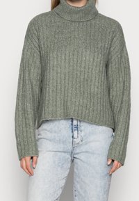 Grön rippad polotröja med en kortad design och långa ärmar, tillverkad av ett strukturerat stickat material, kombinerad med ljusblå jeans.