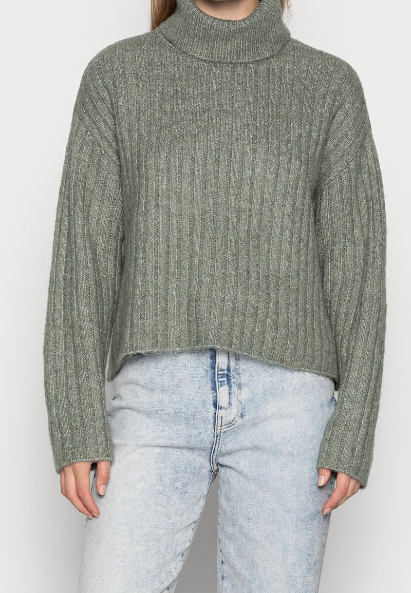 Grön rippad polotröja med en kortad design och långa ärmar, tillverkad av ett strukturerat stickat material, kombinerad med ljusblå jeans.