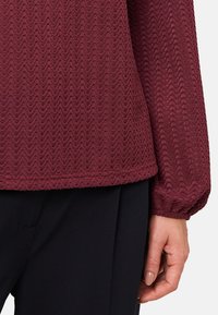 Haut texturé bordeaux avec un motif ondulé, doté de manches bouffantes et d'une coupe décontractée. Assorti à un pantalon noir ajusté.