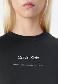 Czarna bawełniana koszulka z okrągłym dekoltem; na przodzie umieszczony biały napis: "Calvin Klein" oraz "ZAŁOŻONA W DZIEWIĘTNASTYM SZEŚĆDZIESIĄTYM ÓSMYM".