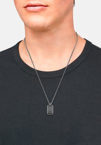Collar metálico de perro con una superficie texturizada, suspendido de una cadena de plata, llevado sobre una camiseta negra de cuello redondo.