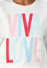 Balta ilgomis rankovėmis marškiniai su ryškiu, daugiakoloriniu tekstu: „VIVA LOVE“ raudonomis, rožinėmis ir mėlynomis raidėmis. Minkšta medžiaga, laisvas siluetas.