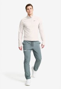 Ljusgrå långärmad zip-up topp i kombination med teal joggerbyxor. Byxorna har en logotyp och båda är tillverkade av lätta, andningsbara material. Vita sneakers.