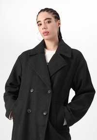 Cappotto nero doppiopetto, realizzato in tessuto spesso, con colletto a risvolto, vestibilità oversize e bottoni grandi. Adatto per il freddo.
