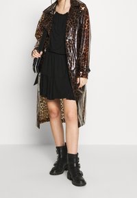 Osoba ubrana w błyszczący płaszcz w cętki lamparta na czarnej sukience, trzymając czarną torbę, i nosząc czarne botki za kostkę z klamrami.