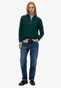 Groene zip-up trui met geribbelde mouwen, over een gestreepte blouse heen gedragen. Lichtblauwe spijkerbroek en donkere schoenen maken de look af.