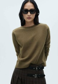 Olivgrüner Cashmere-Pullover mit Rundhalsausschnitt und langen Ärmeln, kombiniert mit hoch geschnittenen Karohosen mit doppelten Schnallenakzenten.