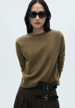 Olivgrüner Cashmere-Pullover mit Rundhalsausschnitt und langen Ärmeln, kombiniert mit hoch geschnittenen Karohosen mit doppelten Schnallenakzenten.