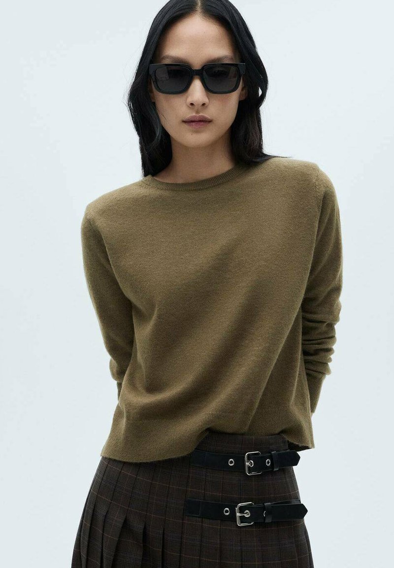 Olivgrüner Cashmere-Pullover mit Rundhalsausschnitt und langen Ärmeln, kombiniert mit hoch geschnittenen Karohosen mit doppelten Schnallenakzenten.
