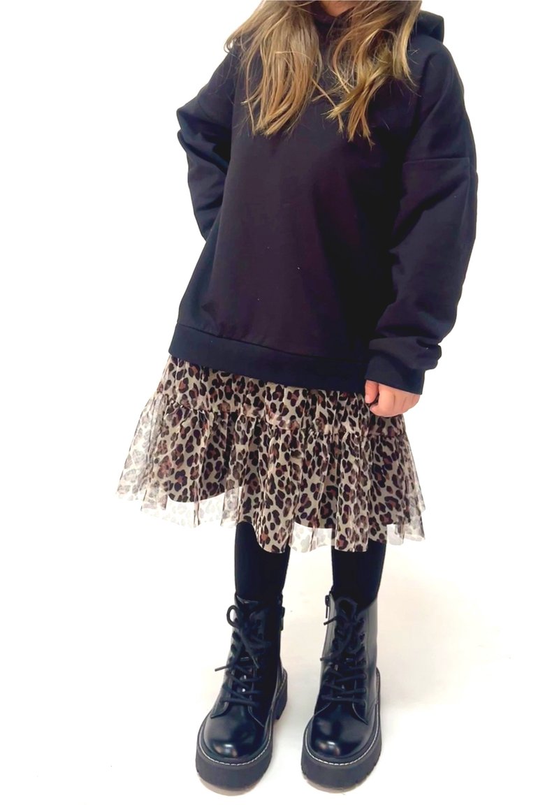 Enfant portant un sweat-shirt noir surdimensionné, une jupe à imprimé léopard avec tulle, des leggings noirs et des bottes noires à lacets, debout devant un fond blanc.