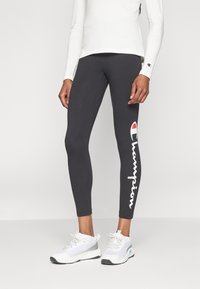 Svarta leggings i elastiskt tyg med en vit "Champion"-logotyp längs sidan, kombinerat med vita träningsskor.