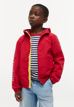 Dreng iført en rød jakke med gul foring over en navy- og hvidstribet skjorte og lyseblå jeans, hænderne i lommerne, ser til siden.