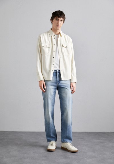Camisa de botones color crema claro sobre una camiseta blanca, combinada con jeans azul claro y zapatillas color blanco roto. Materiales suaves con un ajuste relajado.