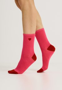 Calcetines de rayas rosas con acentos rojos en la puntera y el talón, que presentan un pequeño diseño de corazón rojo en el lateral. Textura suave y altura hasta la mitad de la pantorrilla.