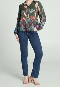 Blusa floreale verde con maniche lunghe, bottoni frontali e vivaci motivi a foglia; abbinata a jeans blu scuro e sandali con tacco a blocco color nudo.