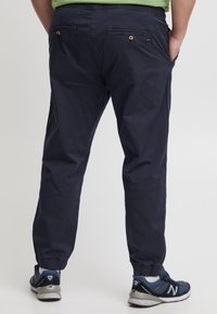 Marine joggpants met elastische boorden, voorzien van achterzakken met knoopsluitingen en een relaxed fit, gedragen met blauwe sportschoenen.