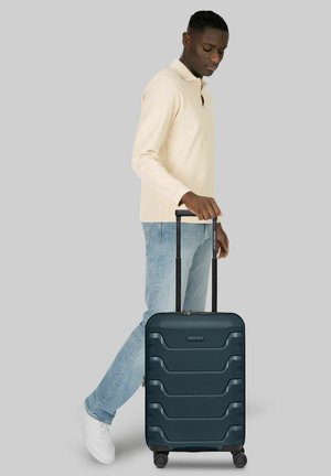 Uomo con camicia beige a maniche lunghe e jeans che cammina trascinando un trolley rigido nero con manico esteso.