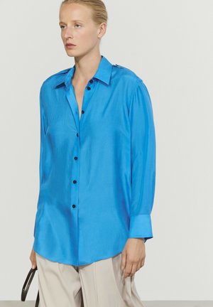 Donna che indossa una camicia blu brillante a maniche lunghe con bottoni e pantaloni beige chiaro, tenendo una cinghia nera con uno sfondo neutro.