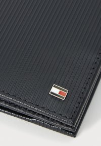 Zwarte geribbelde portemonnee van leer. Beschikt over een klein rechthoekig metalen logo met rode, witte en blauwe accenten. Compacter en gestructureerd ontwerp.