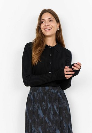 Zwarte geribbelde top met lange mouwen, een kraag en een knoopsluiting aan de voorkant; beschikt over duimgaten. Gekleed met een patronenrok in donkerblauw en zwart.