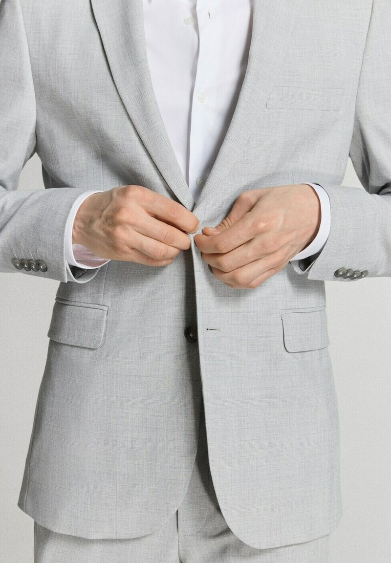 Man die een lichtgrijs pak en wit overhemd draagt en zijn jasje dichtknopen, close-up vanaf de taille gezien.