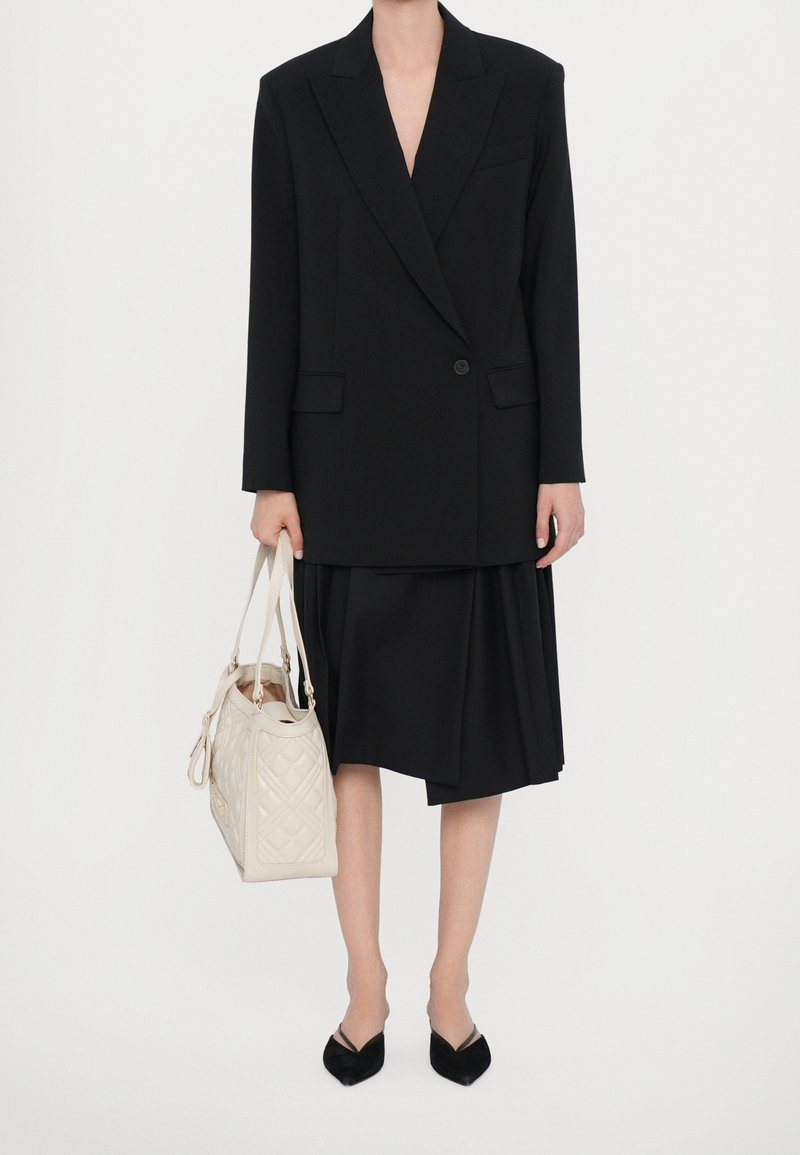 Blazer noir oversize avec larges revers, associé à une jupe asymétrique noire. Portant un sac à main en cuir beige matelassé, avec des ballerines noires à bouts pointus.