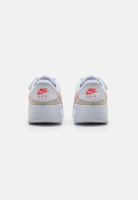 Nike Air sportbačiai su balta tinkline viršutine dalimi, šviesiai rožinėmis detalėmis ir tekstūruota pilka padais. Logotipas aiškiai matomas raudonai.